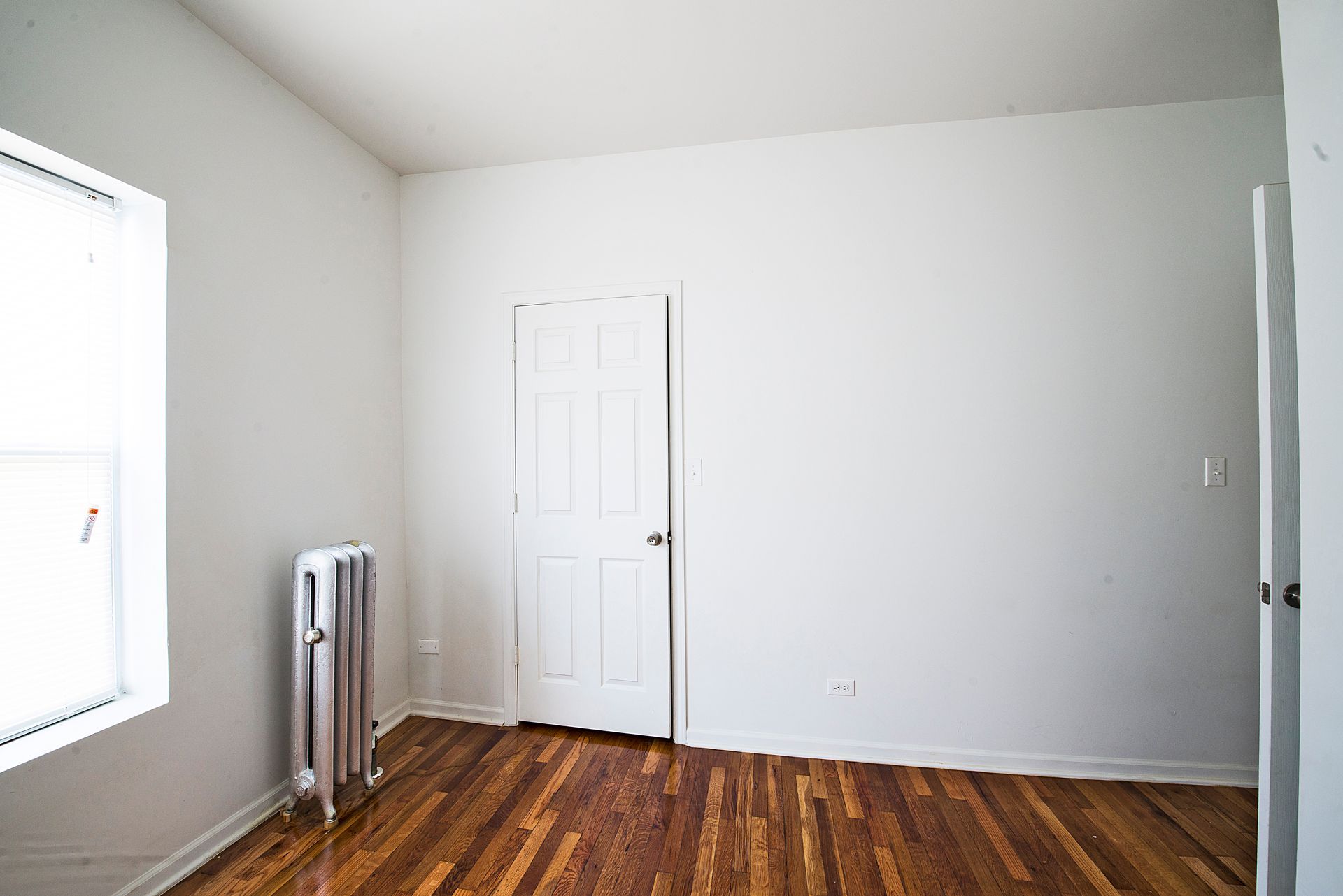 Empty room with a window, white walls, wooden floor, and a door. There is a radiator on the wall.