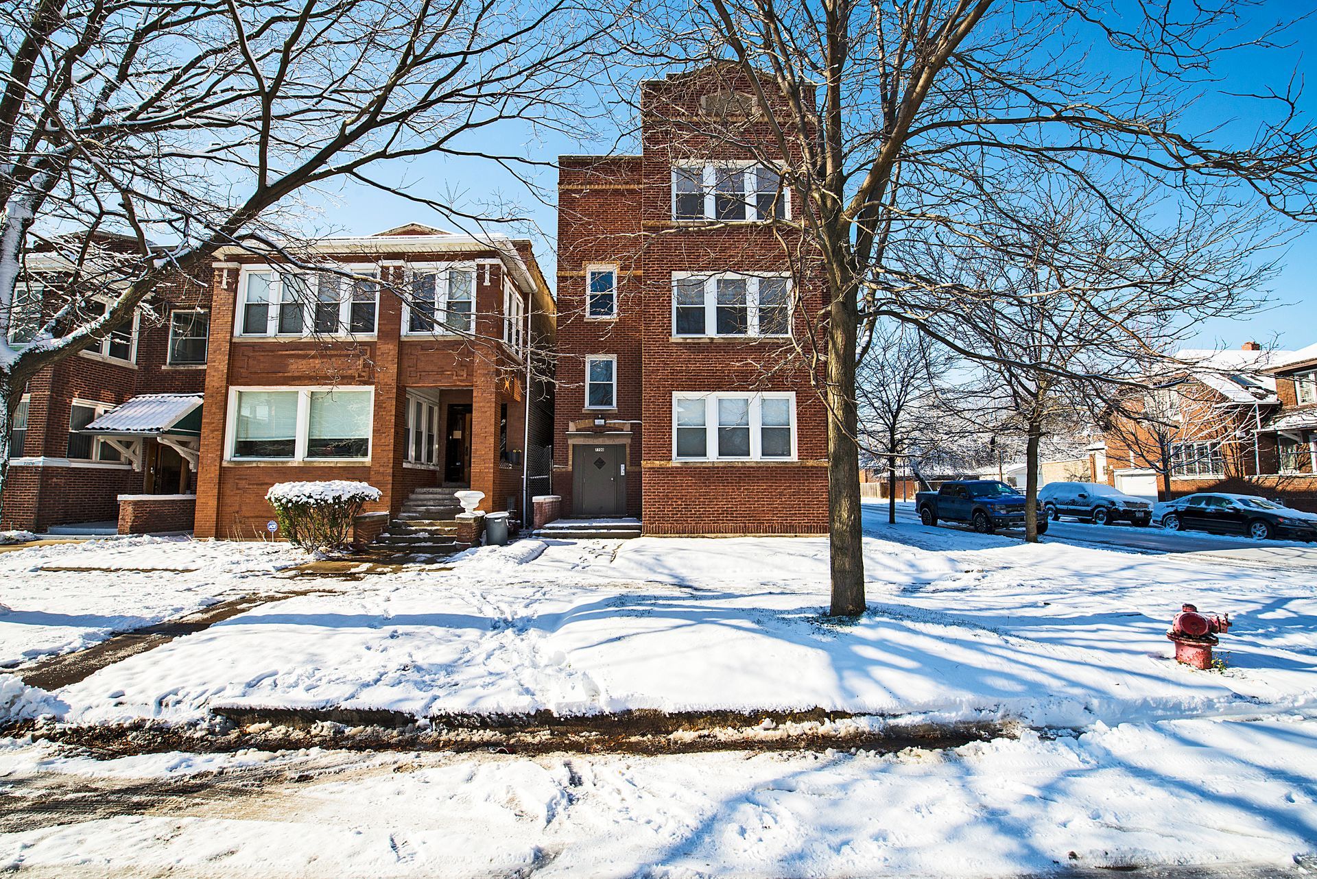 Snow-covered brick buildings on a sunny winter day, with trees and a fire hydrant in the foreground.