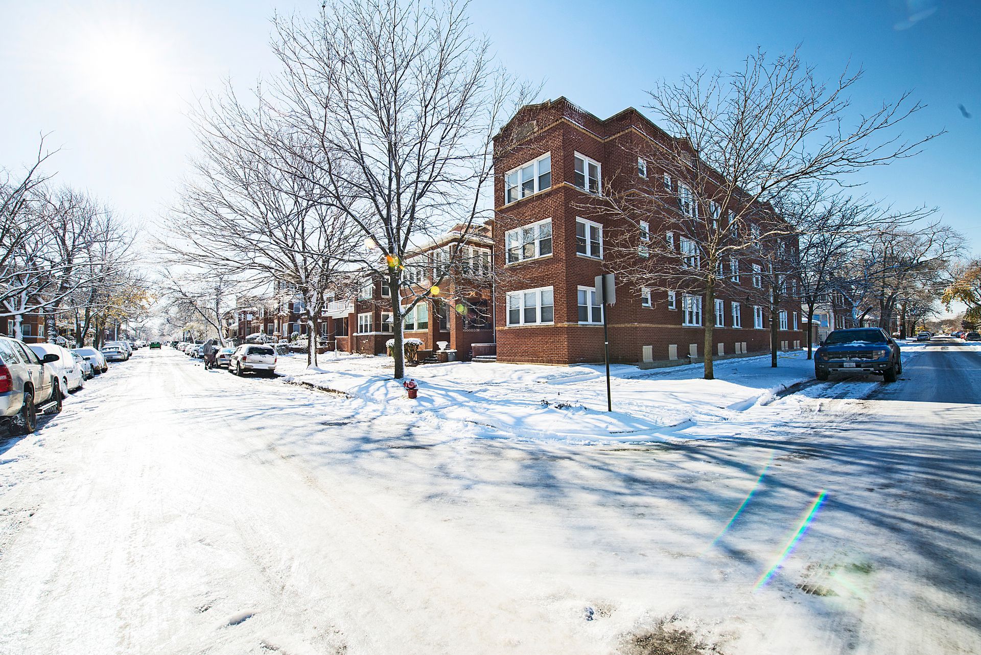 Snow-covered street in front of a brick apartment building; cars parked along the street, trees bare.