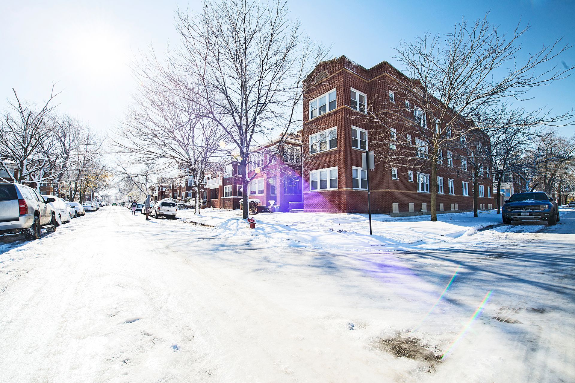 Snowy street with brick apartment building; cars parked along the sides, trees, and bright sun.