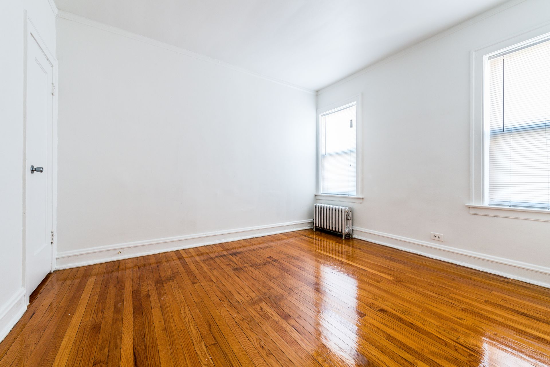 Empty room with hardwood floors, white walls, two windows with blinds, and a radiator.