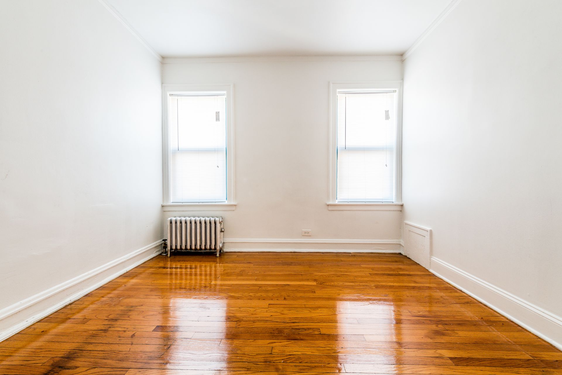 Empty room with hardwood floors, white walls, two windows, and radiator.
