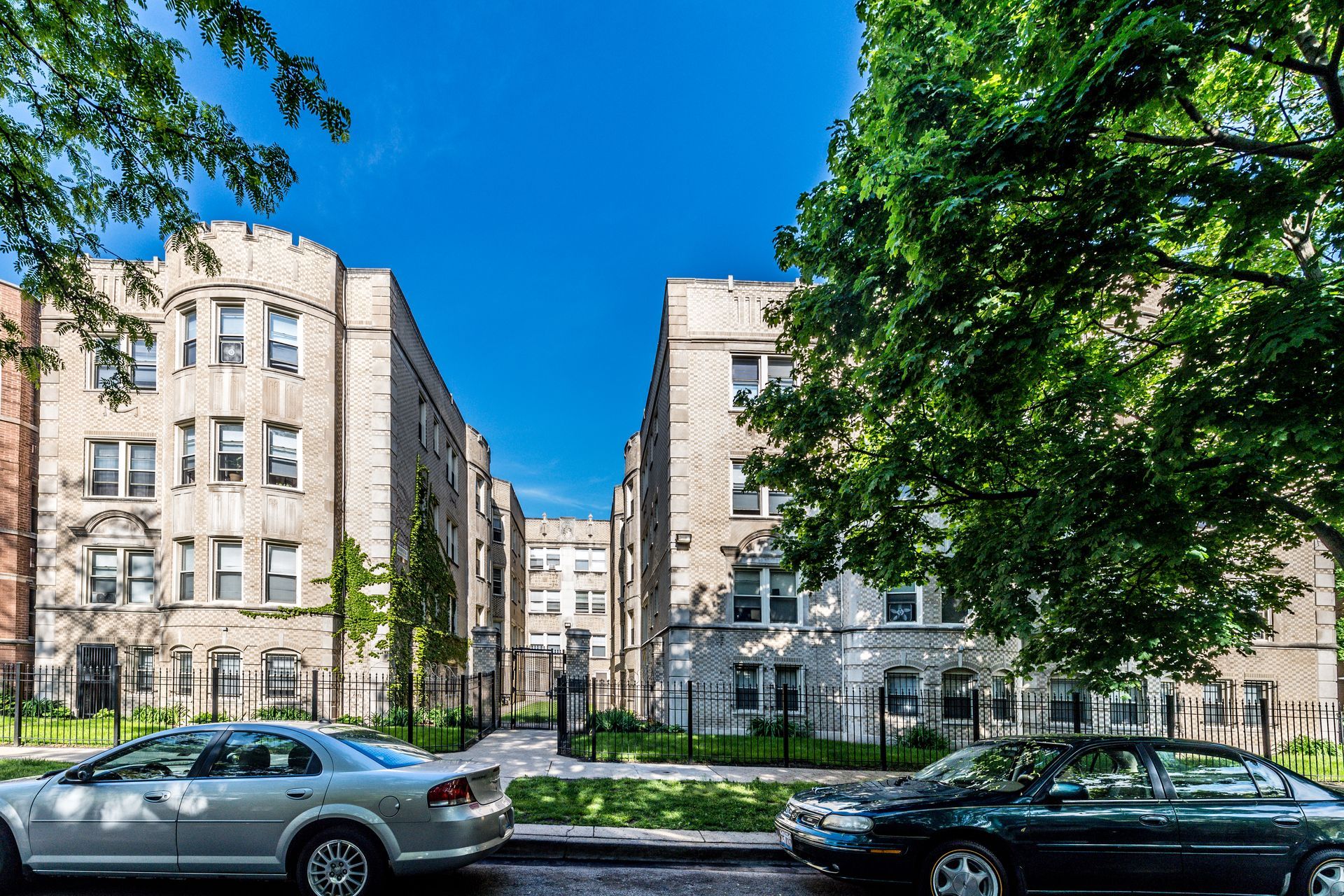 Apartment buildings with arched details, trees, and parked cars under a bright blue sky.