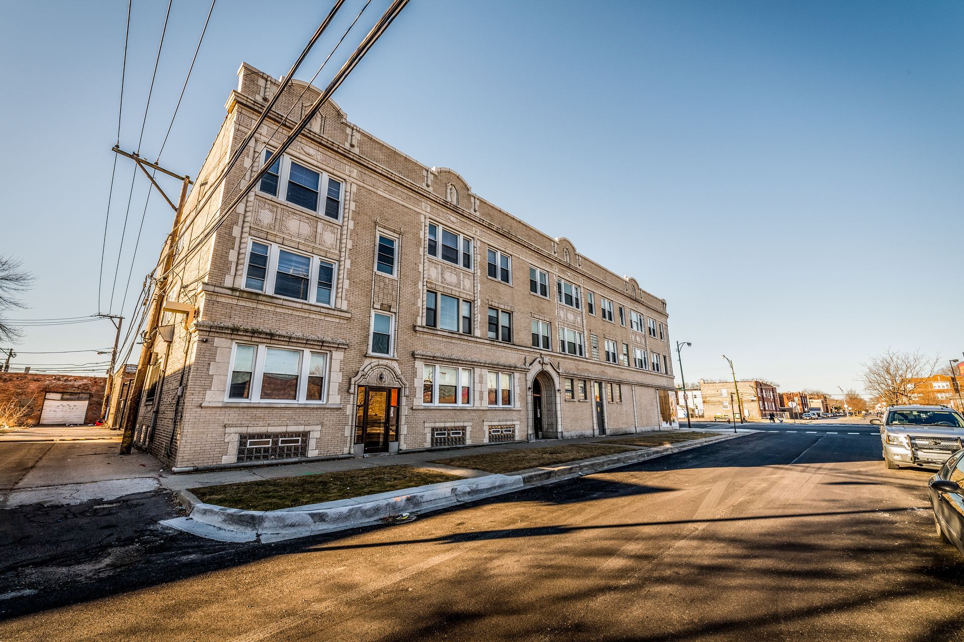 Three-story beige apartment building on a sunny street, utility poles and car visible.