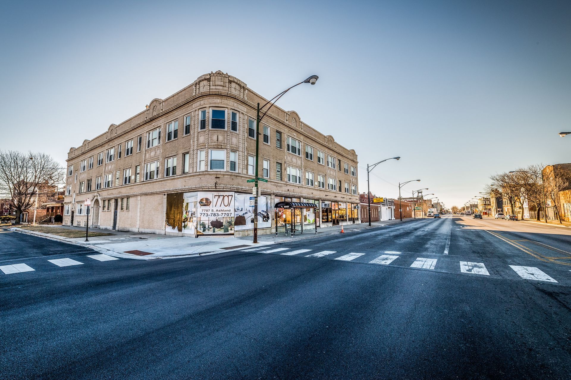 Two-story building at a street corner with crosswalk. Graffiti on lower level. Empty street, clear sky.