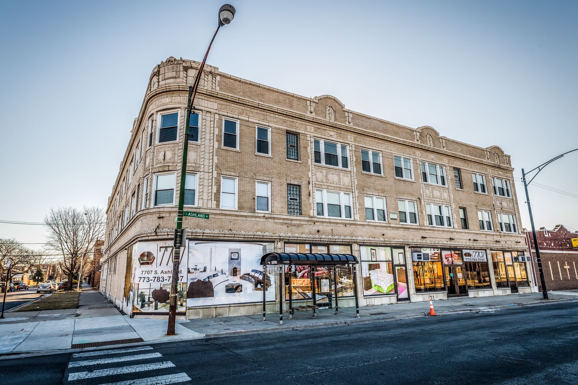 Three-story building with storefronts and apartments. Brown brick facade, street scene, bus stop, and bare trees.