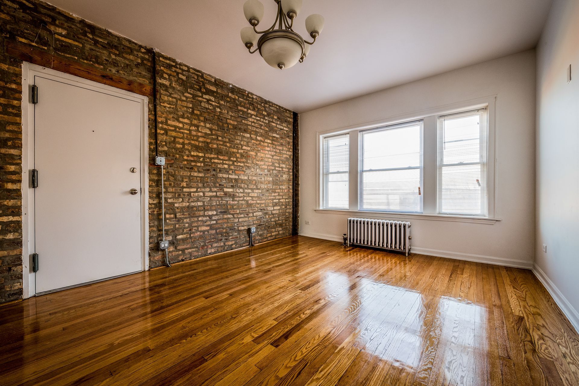 Empty room with hardwood floors, brick wall, white door and window, and a chandelier.