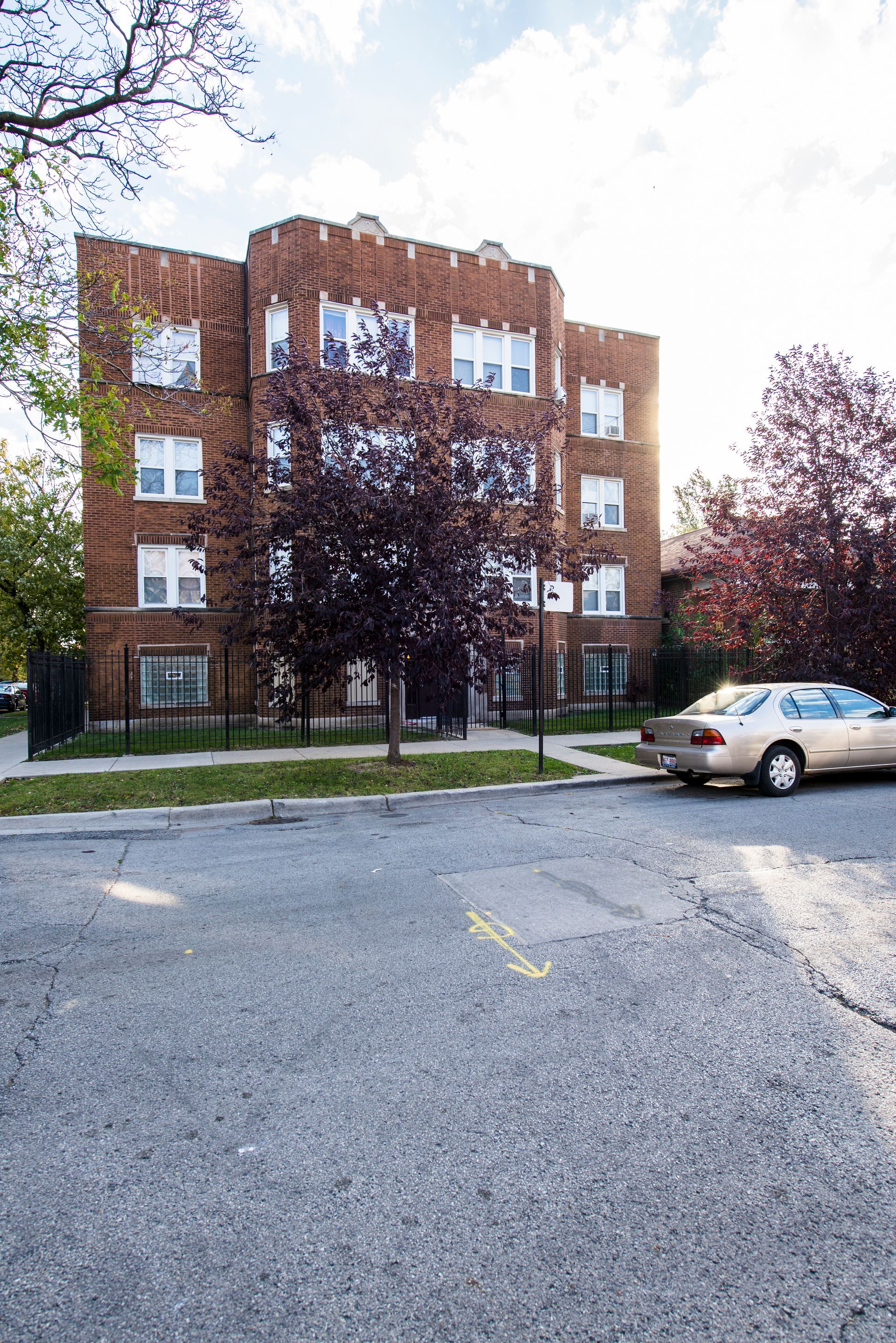 Brick apartment building with a car parked on the street. Trees and a sunny sky are visible.