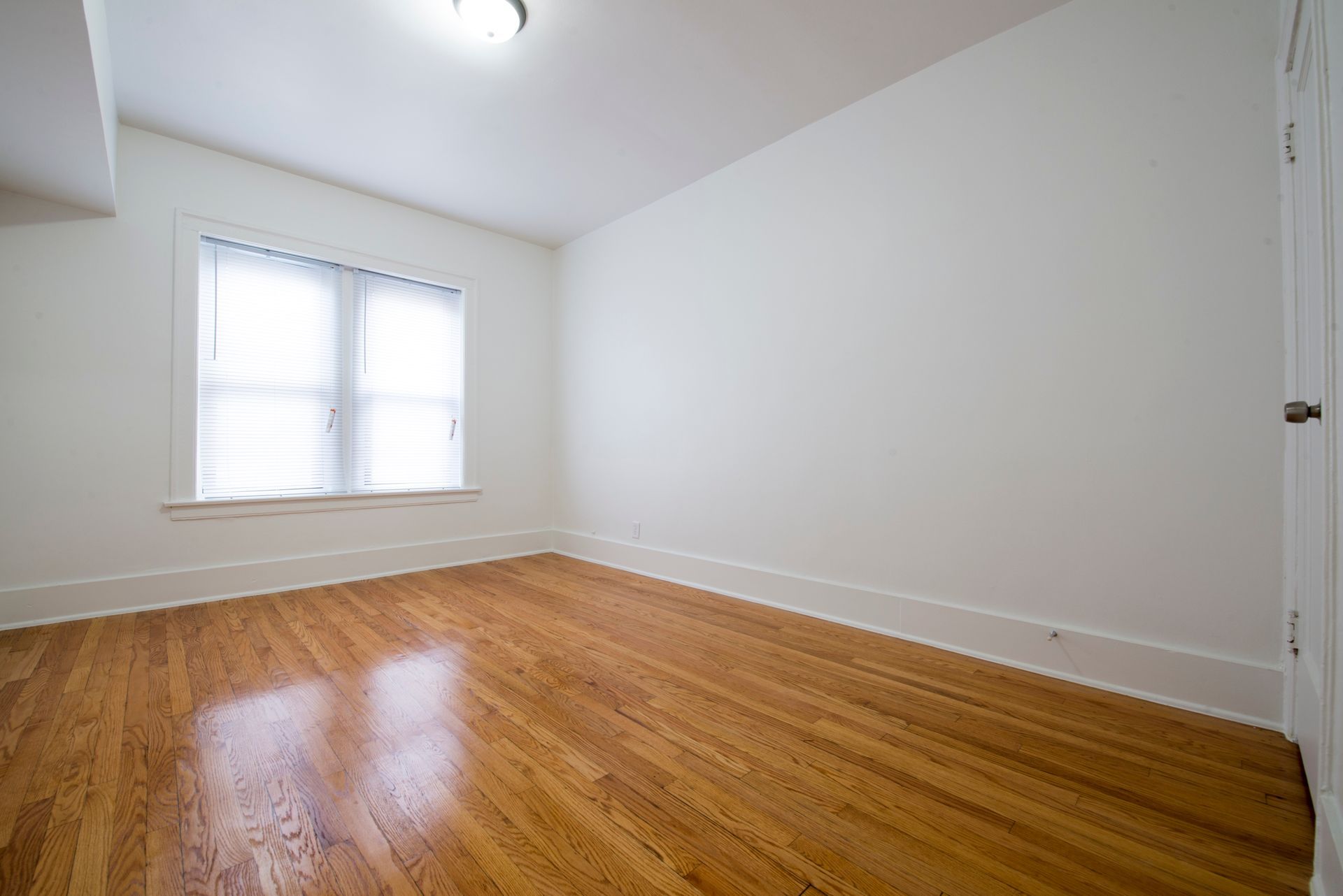 Empty room with hardwood floors, a window with blinds, and white walls.