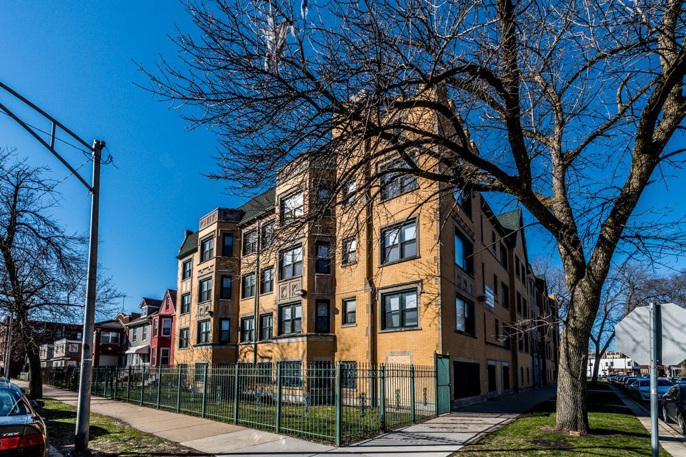 Yellow brick apartment building with dark trim, fence, and tree on a sunny day.