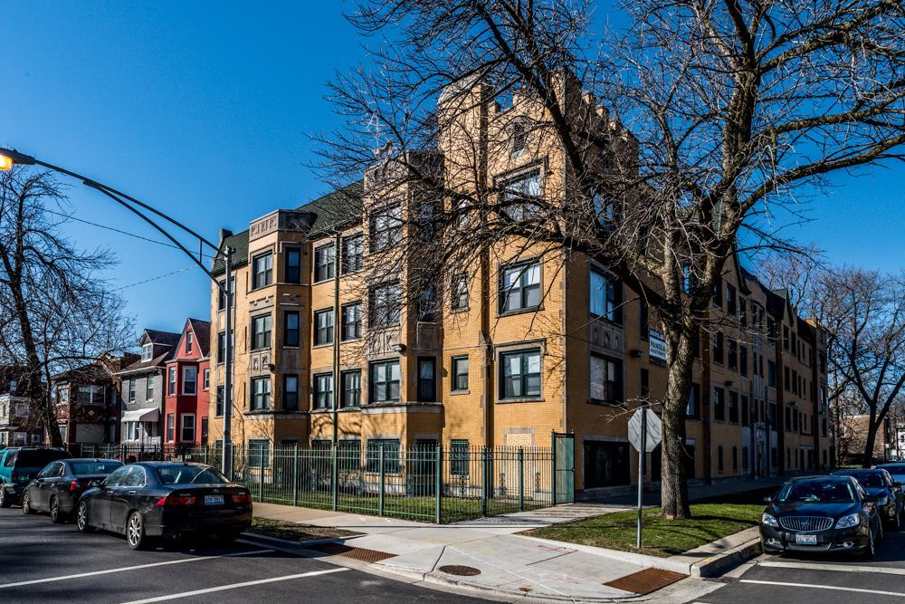 Apartment building on a corner lot; tan brick facade, small fenced yard. Cars parked on street.