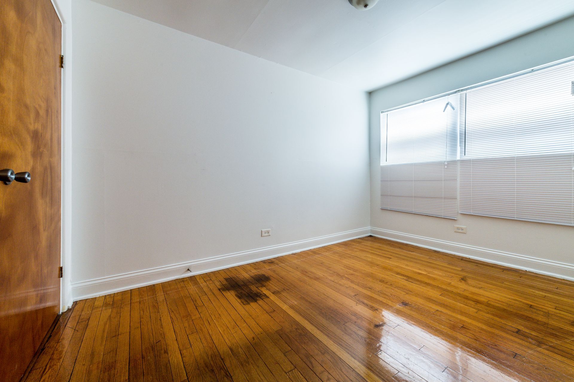 Empty room with wooden floor, white walls, closed door on the left, window with blinds on the right.