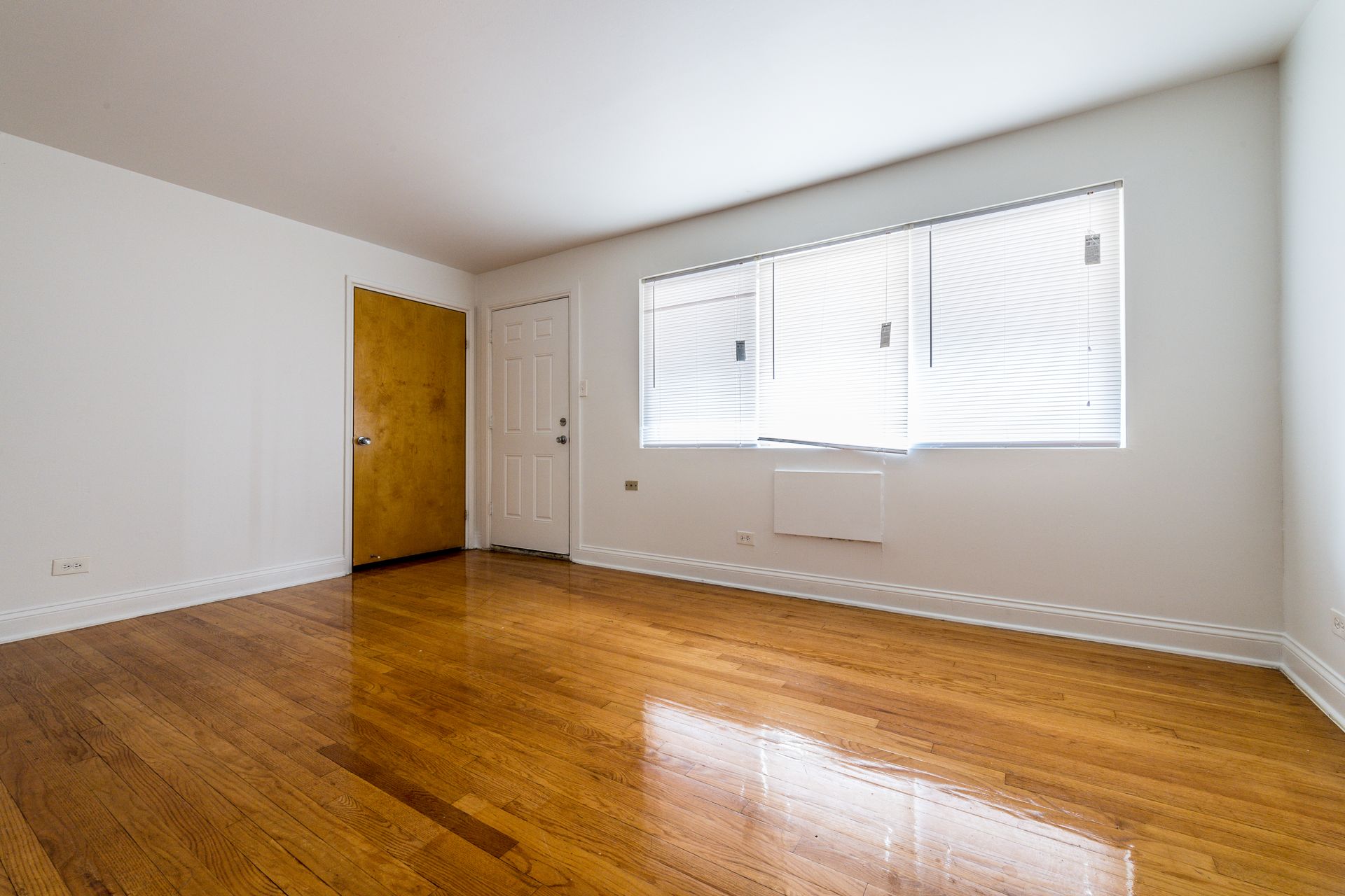Empty room with hardwood floors, white walls, closed door, window with blinds.
