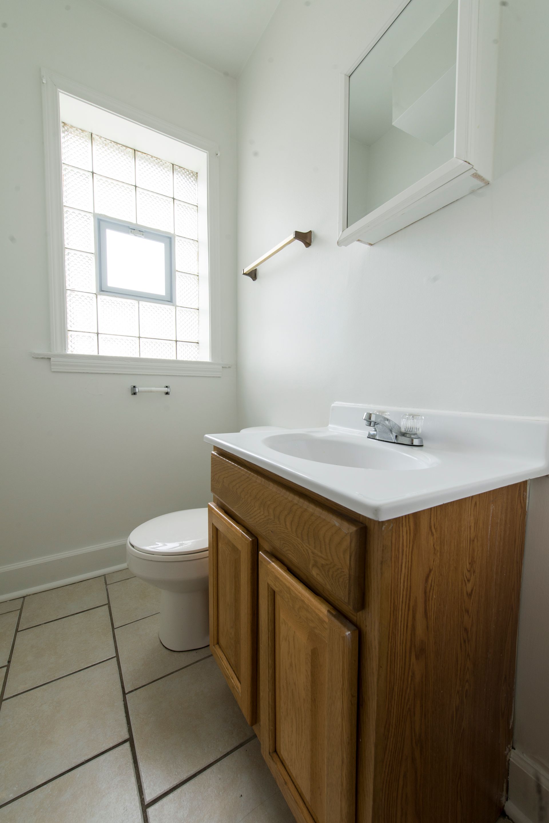 Bathroom with wooden vanity, white toilet, window, and mirror. Beige tiled floor.
