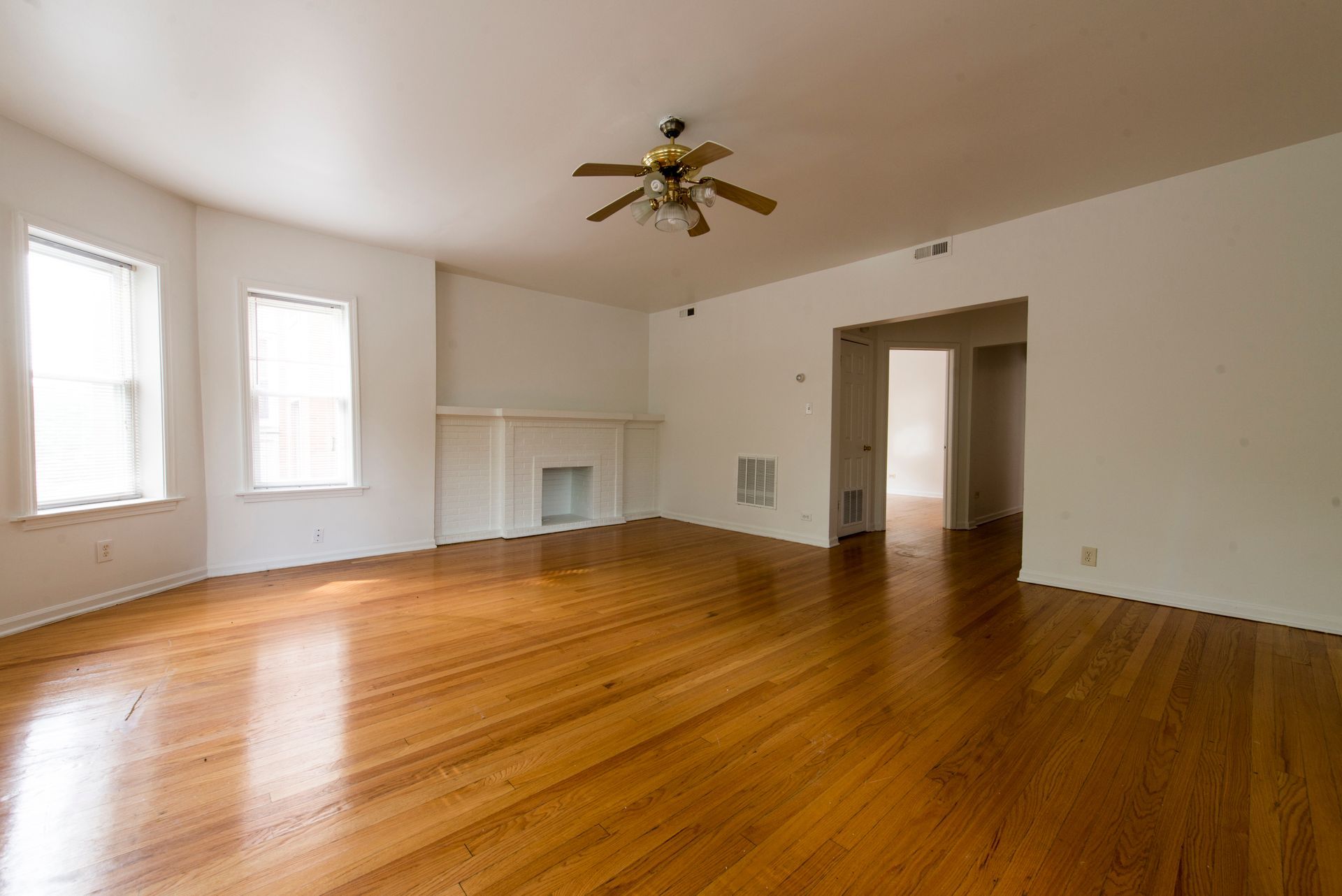 Empty living room with wood floor, white walls, and fireplace. Ceiling fan and doorway to another room.