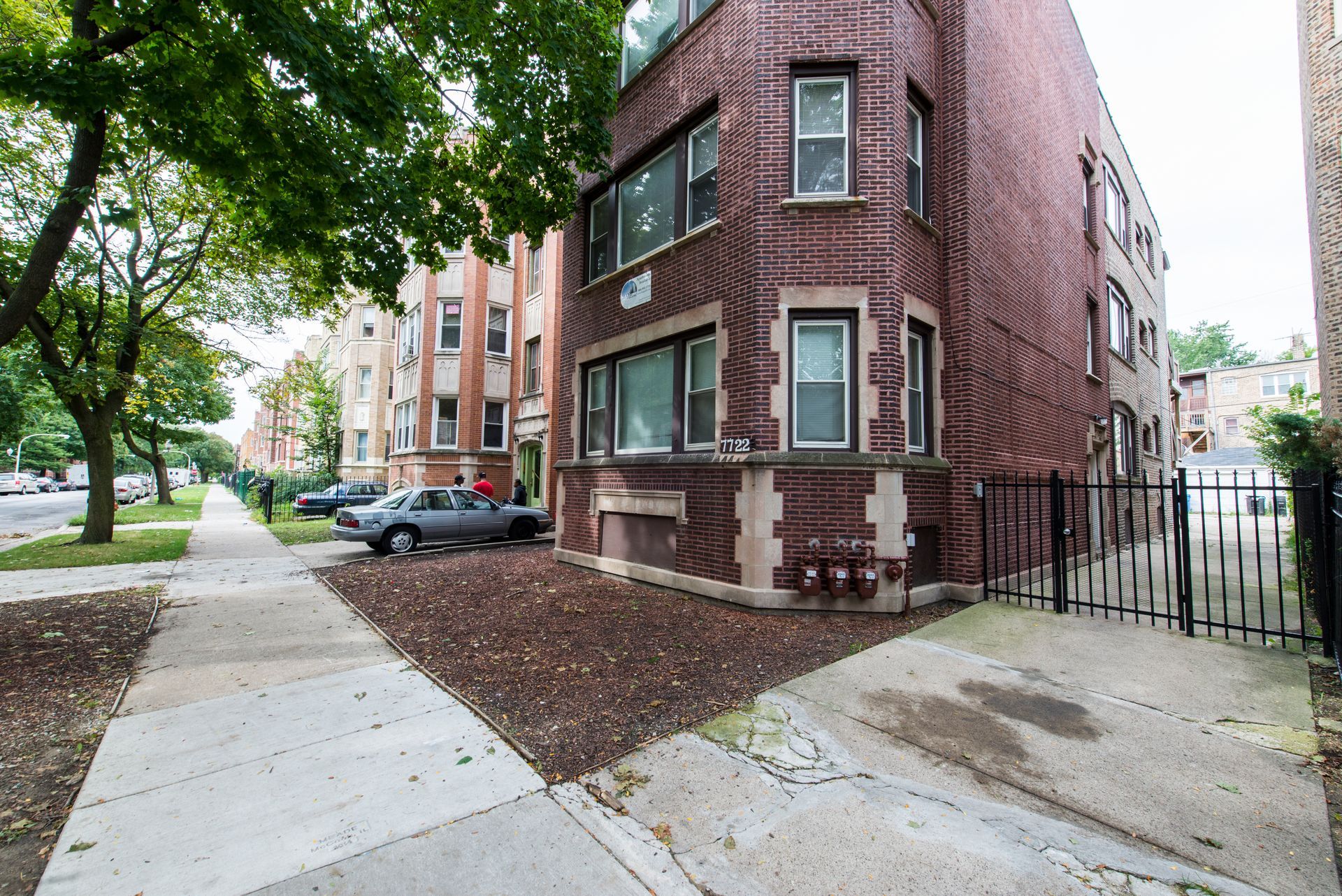 Brick apartment building on a tree-lined street with a sidewalk. Gray car parked in front.