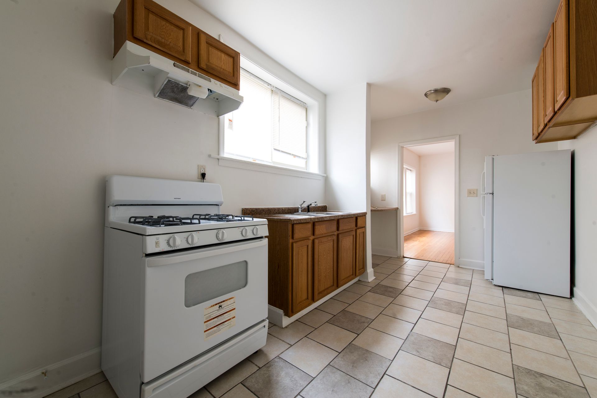 Kitchen with white appliances, wooden cabinets, checkered floor, and a doorway to another room.