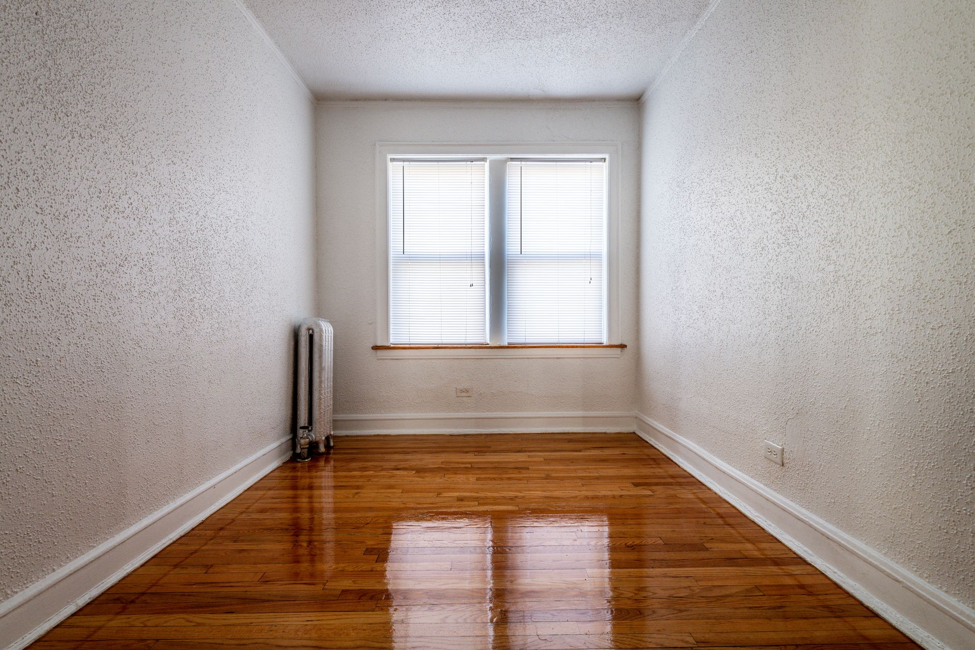 Empty room with hardwood floors, window with blinds, and textured walls.