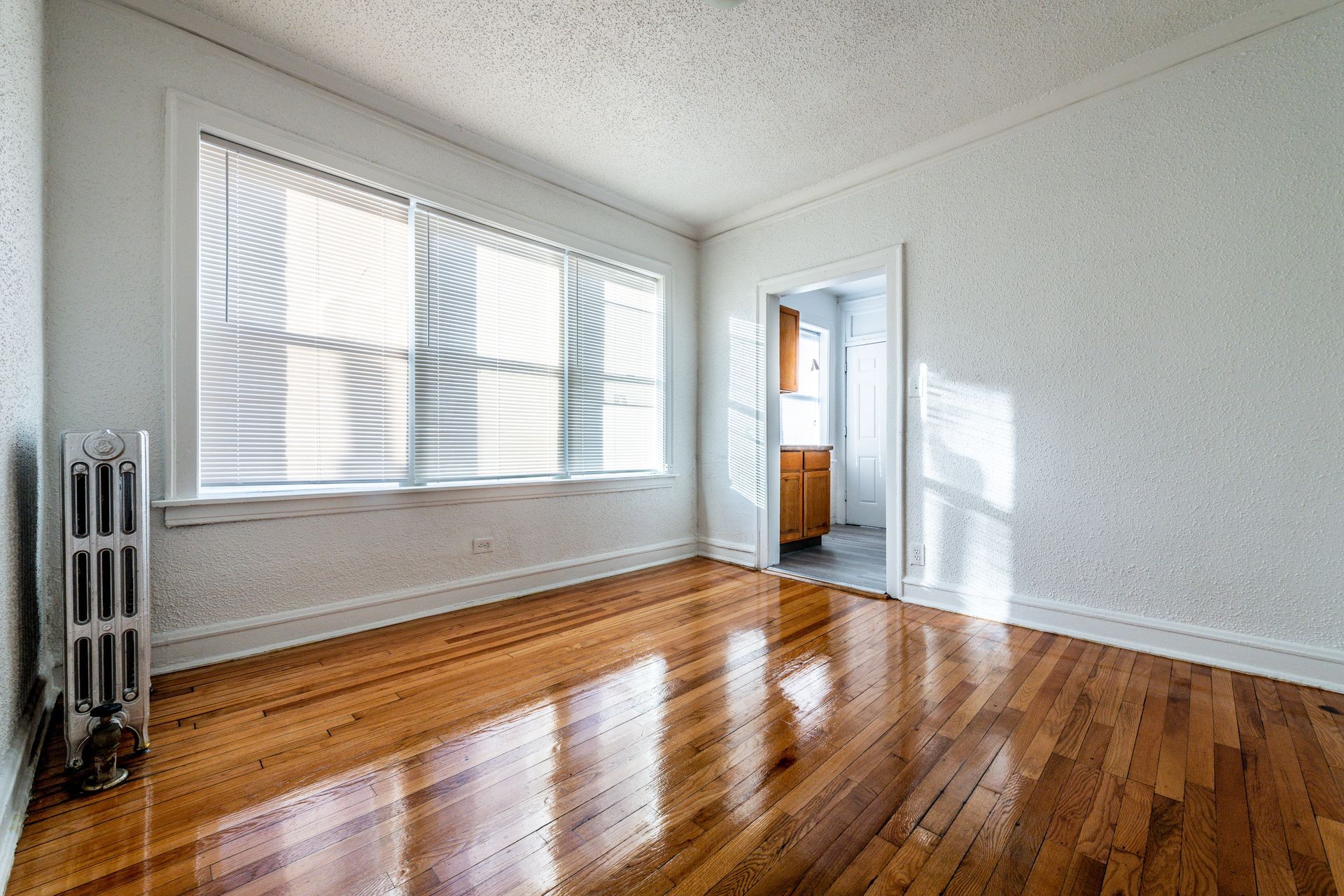Empty room with hardwood floors, a large window, radiator, and a doorway to a kitchen.