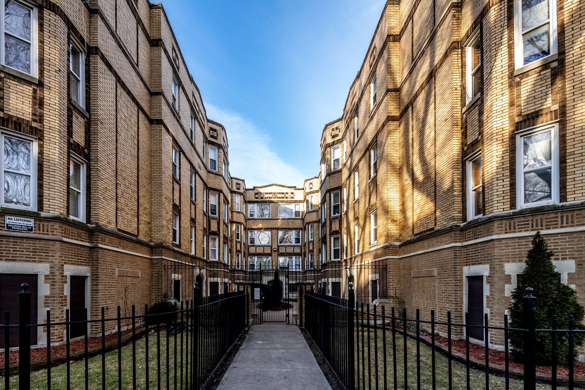 Two brick apartment buildings face each other, connected by a wrought-iron gate and pathway under a blue sky.