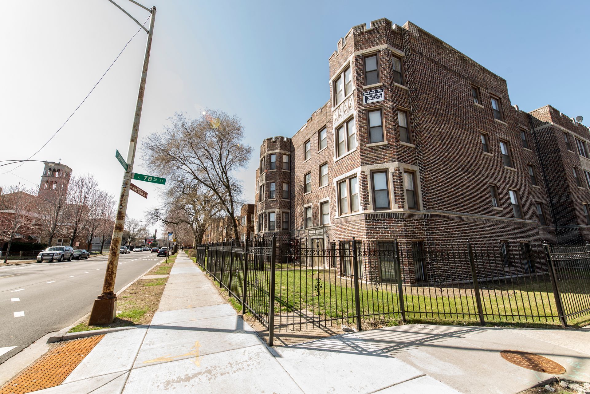 Brick apartment building with turrets, sidewalk, street, and a fence on a sunny day.