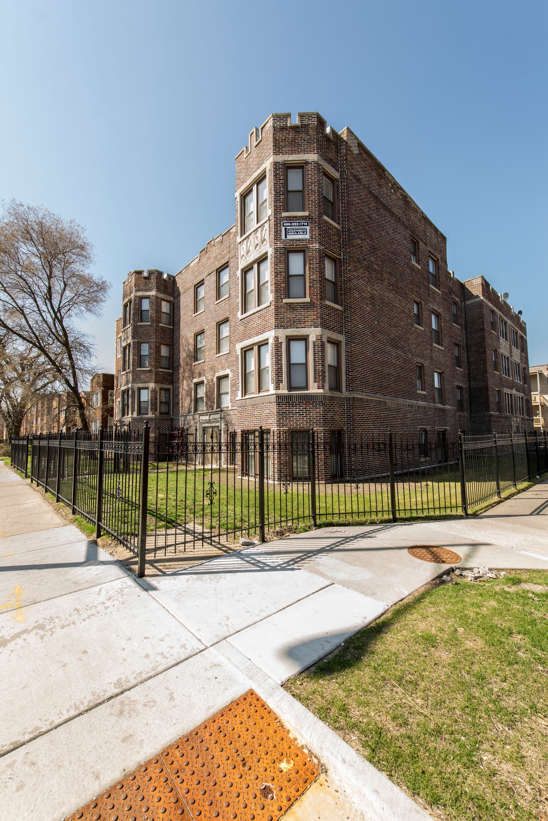 Brick apartment building behind a black fence, on a sunny day. Sidewalk and grass in the foreground.