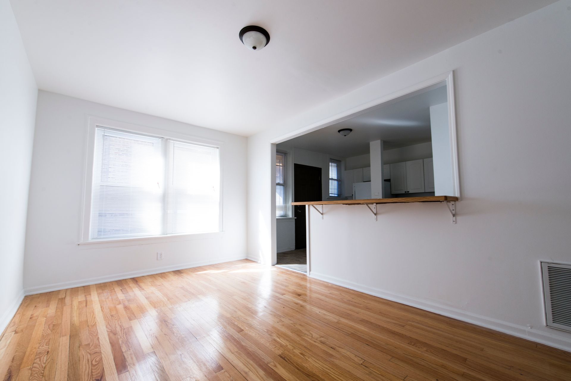 Empty room with hardwood floors, a pass-through to a kitchen, and a window with blinds.