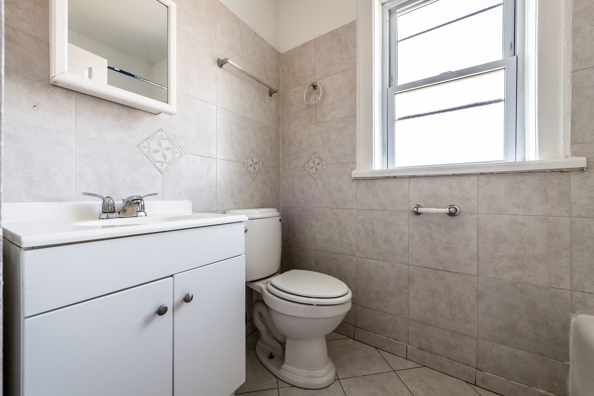Bathroom with white vanity, toilet, and window. Tile walls and floor, and a mirror above the sink.