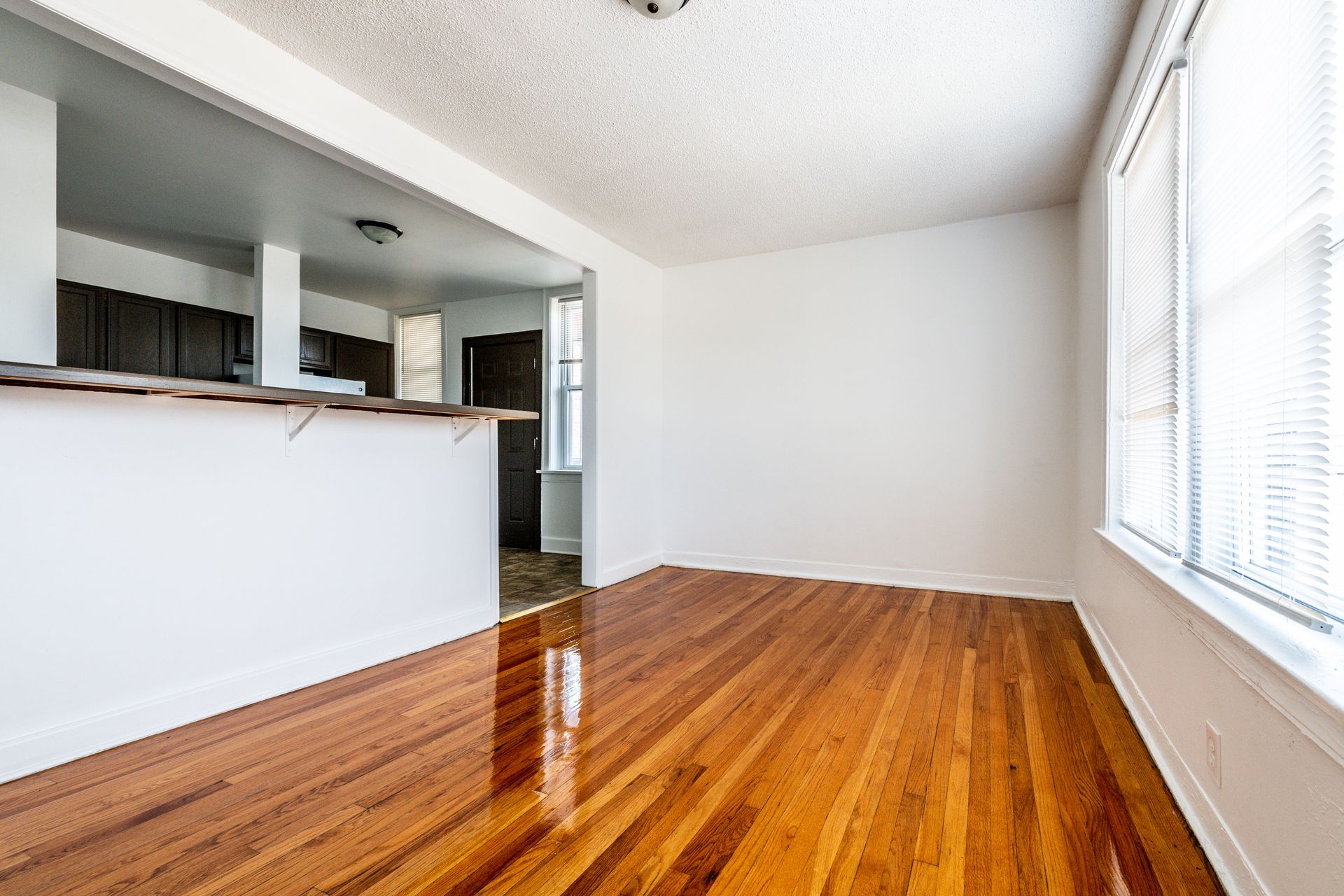 Empty room with hardwood floors, white walls, and a view into a kitchen.