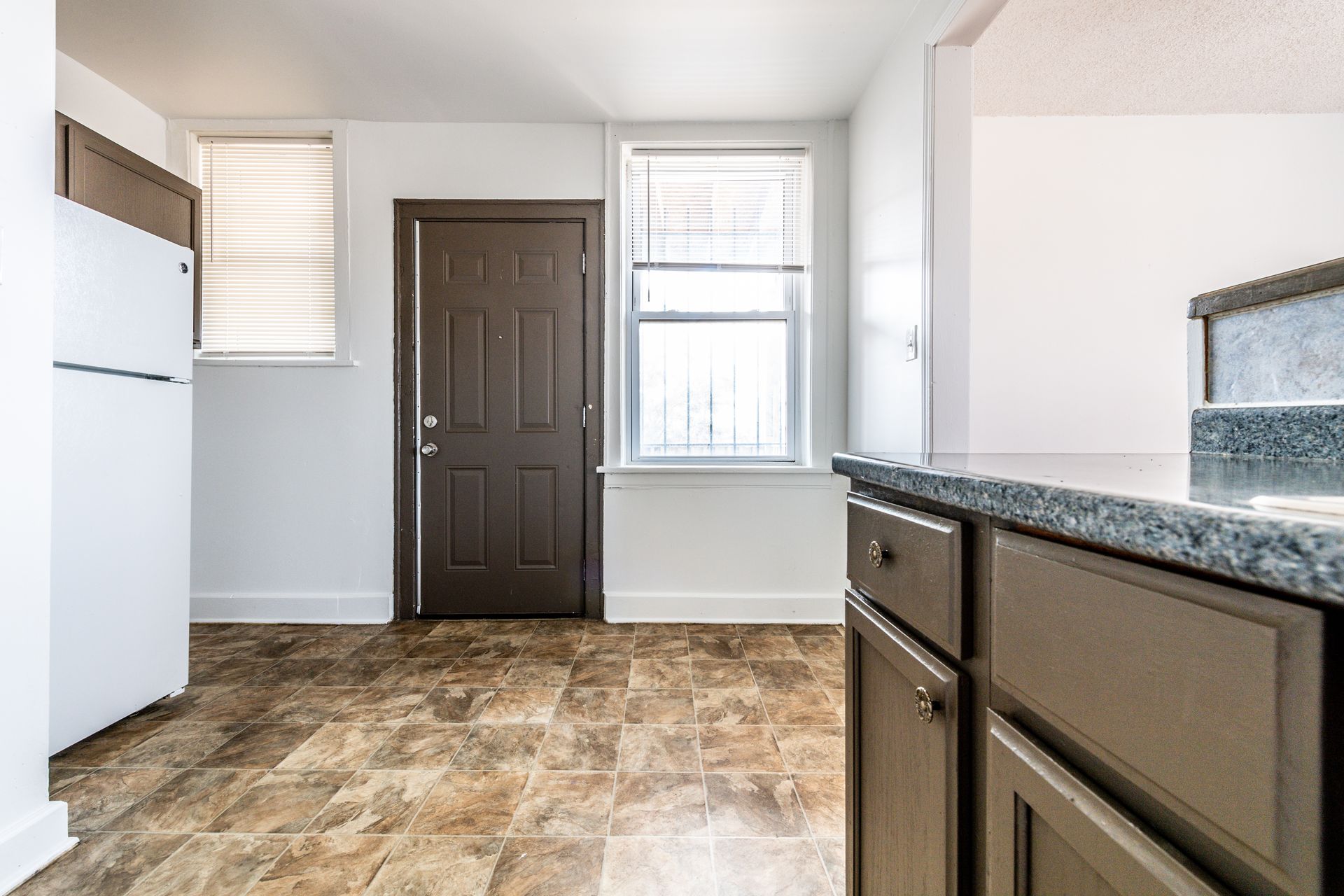 Kitchen with white refrigerator, dark cabinets, brown door, and windows with blinds.