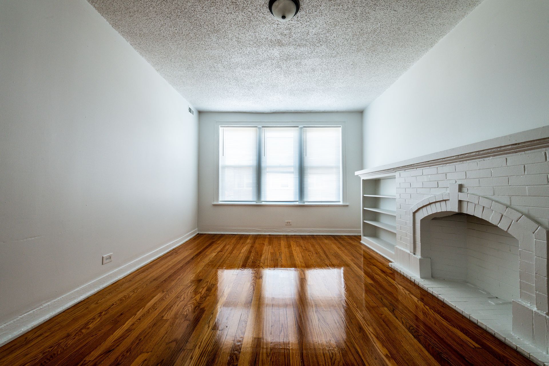 Empty room with hardwood floors, white walls, built-in fireplace, and window with blinds.