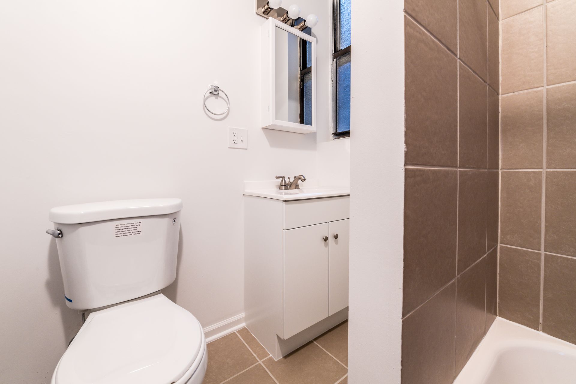 A small bathroom with a toilet, sink, mirror, and a brown tiled shower stall.