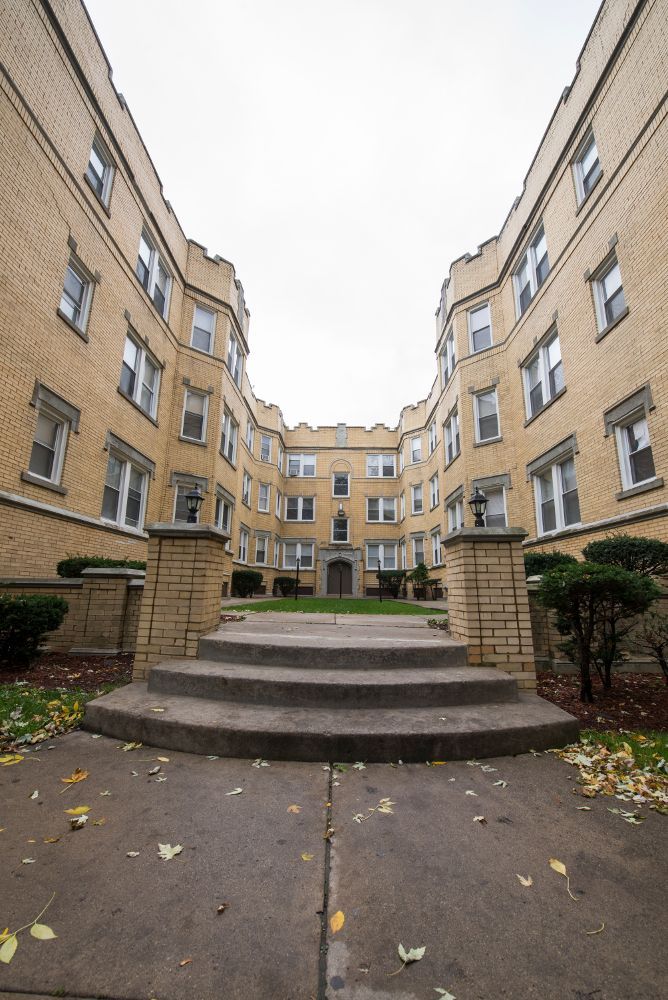 Courtyard apartment building with steps leading toward the entrance; beige brick, overcast sky.