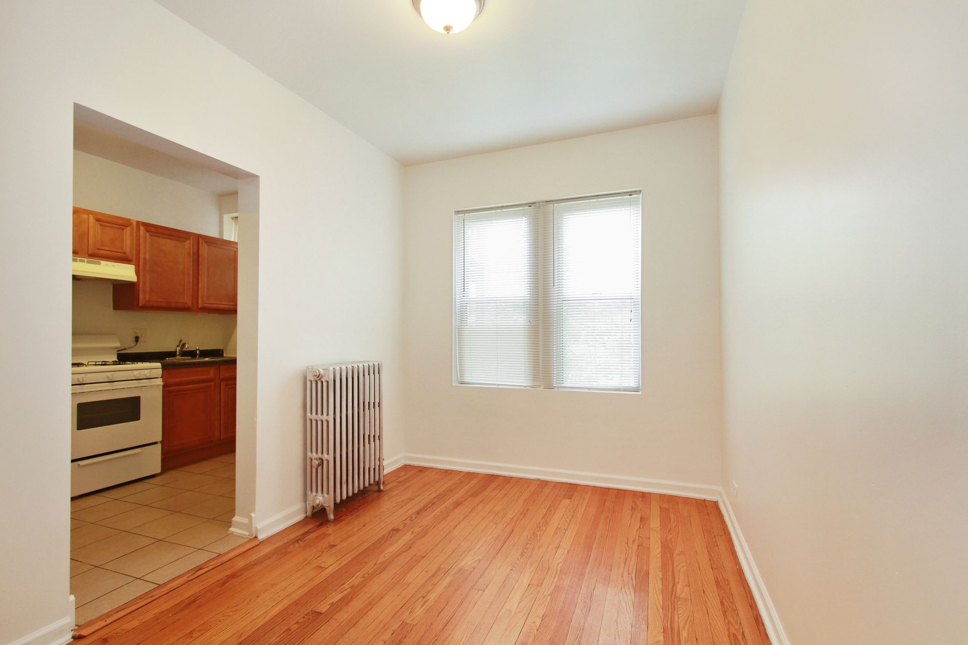 Empty room with wood floors, radiator, and a view into a kitchen.
