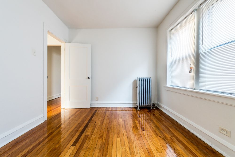 Empty room with hardwood floors, white walls, closed door, radiator, and window with blinds.