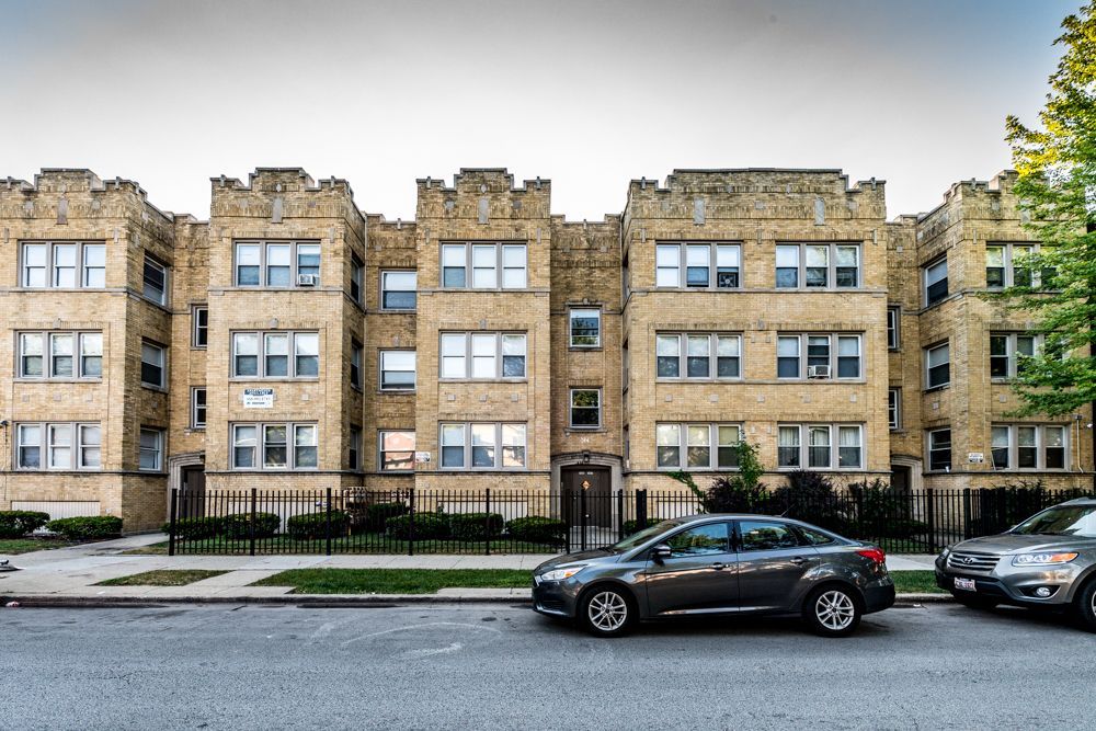 Apartment building with yellow brick facade, small wrought iron fence, cars parked on street.