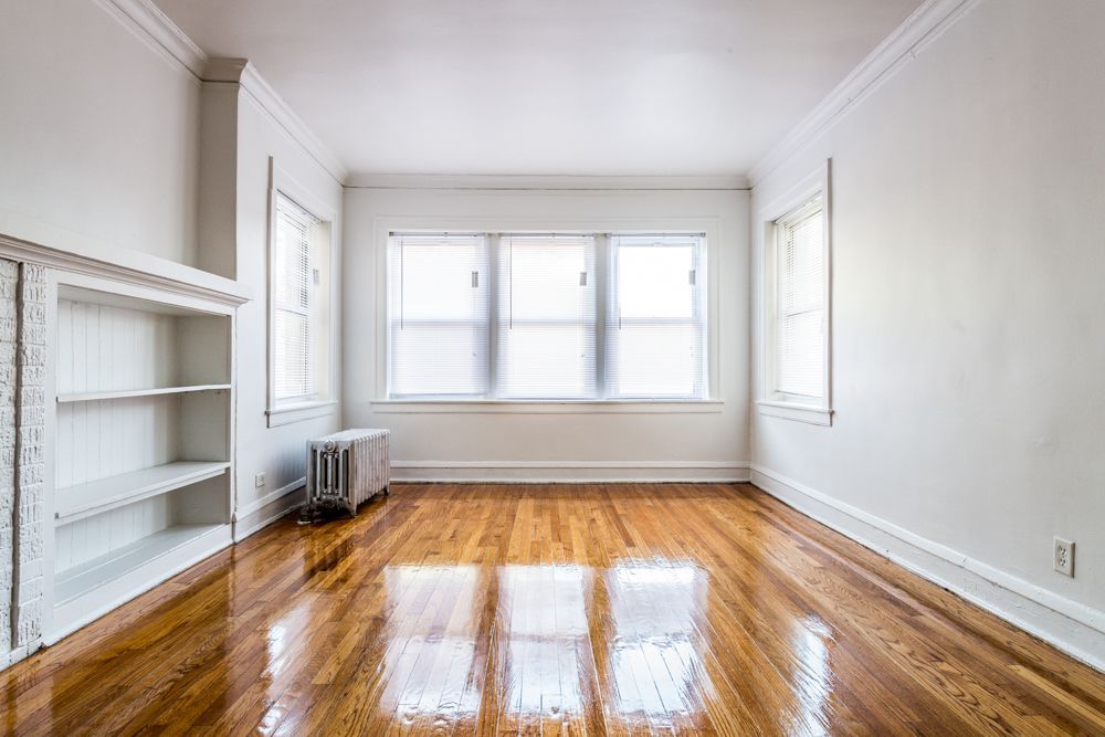 Empty room with glossy hardwood floors, white walls, built-in shelving, and a large window.