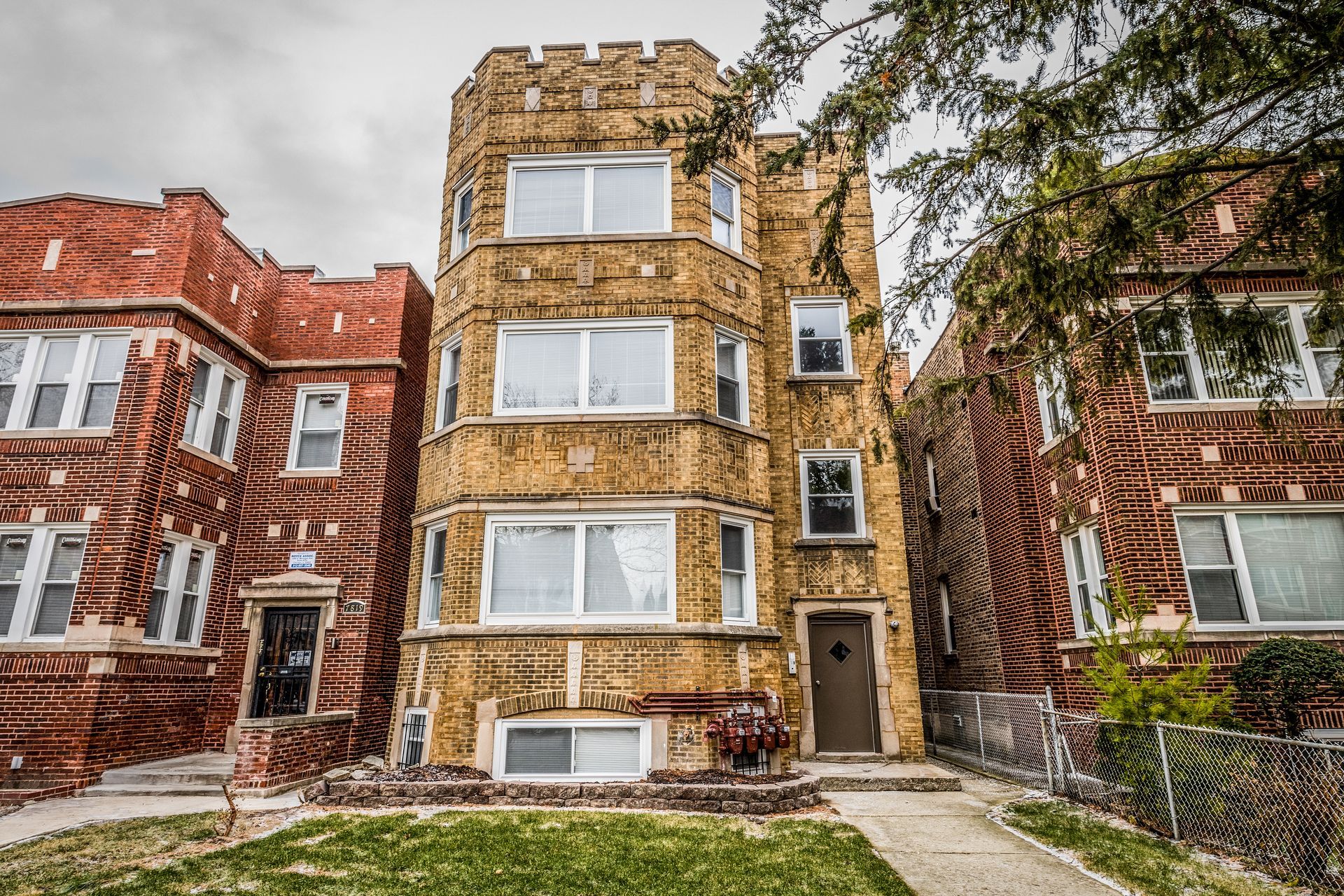 Three-story brick apartment building with a castle-like design between two similar brick buildings, green grass and cloudy sky.