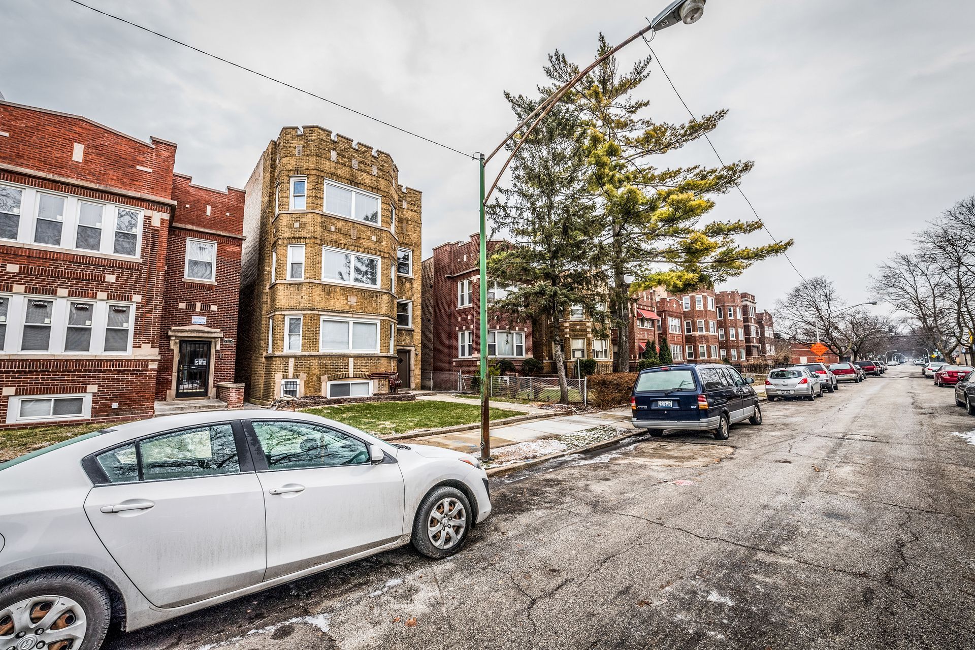 Street with parked cars and brick apartment buildings on a cloudy day.