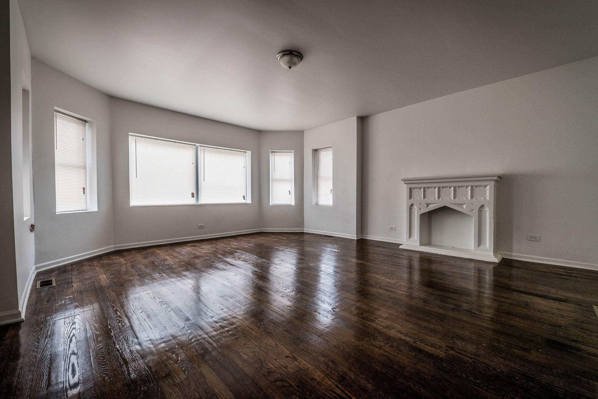 Empty room with dark hardwood floors, white walls, fireplace, and window coverings.
