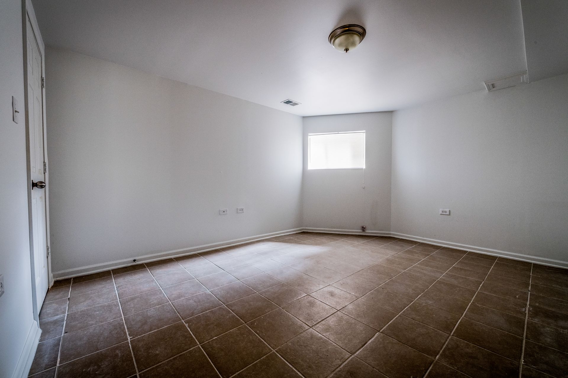 Empty room with brown tiled floor, white walls, small window, and ceiling light.