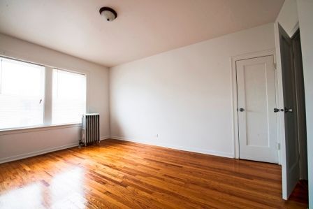 Empty room with hardwood floors, white walls, and a closed door. Window on the left with a radiator.