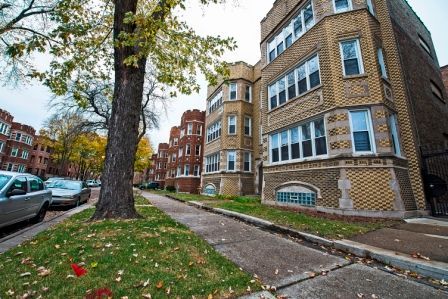 Apartment buildings along a sidewalk with parked cars and a tree in autumn.