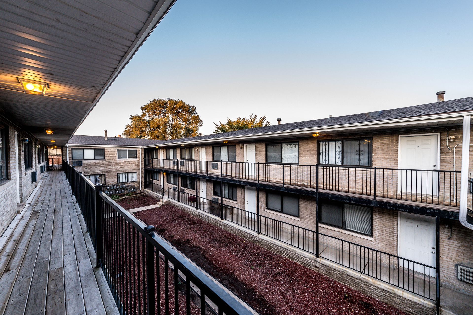 Exterior of two-story apartment building with walkways, black railings, and brick facade. Red mulch fills the inner garden.