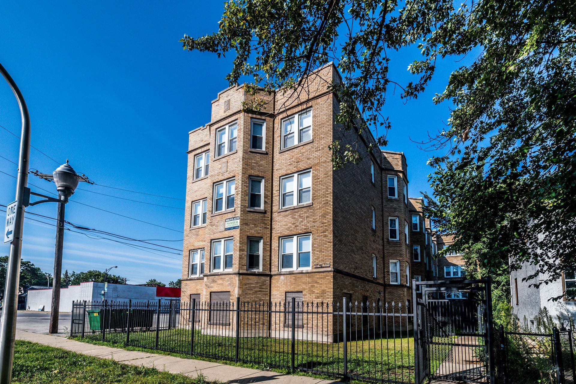 Multi-story brick apartment building on a sunny day. Metal fence in front, tree branches overhead, street.