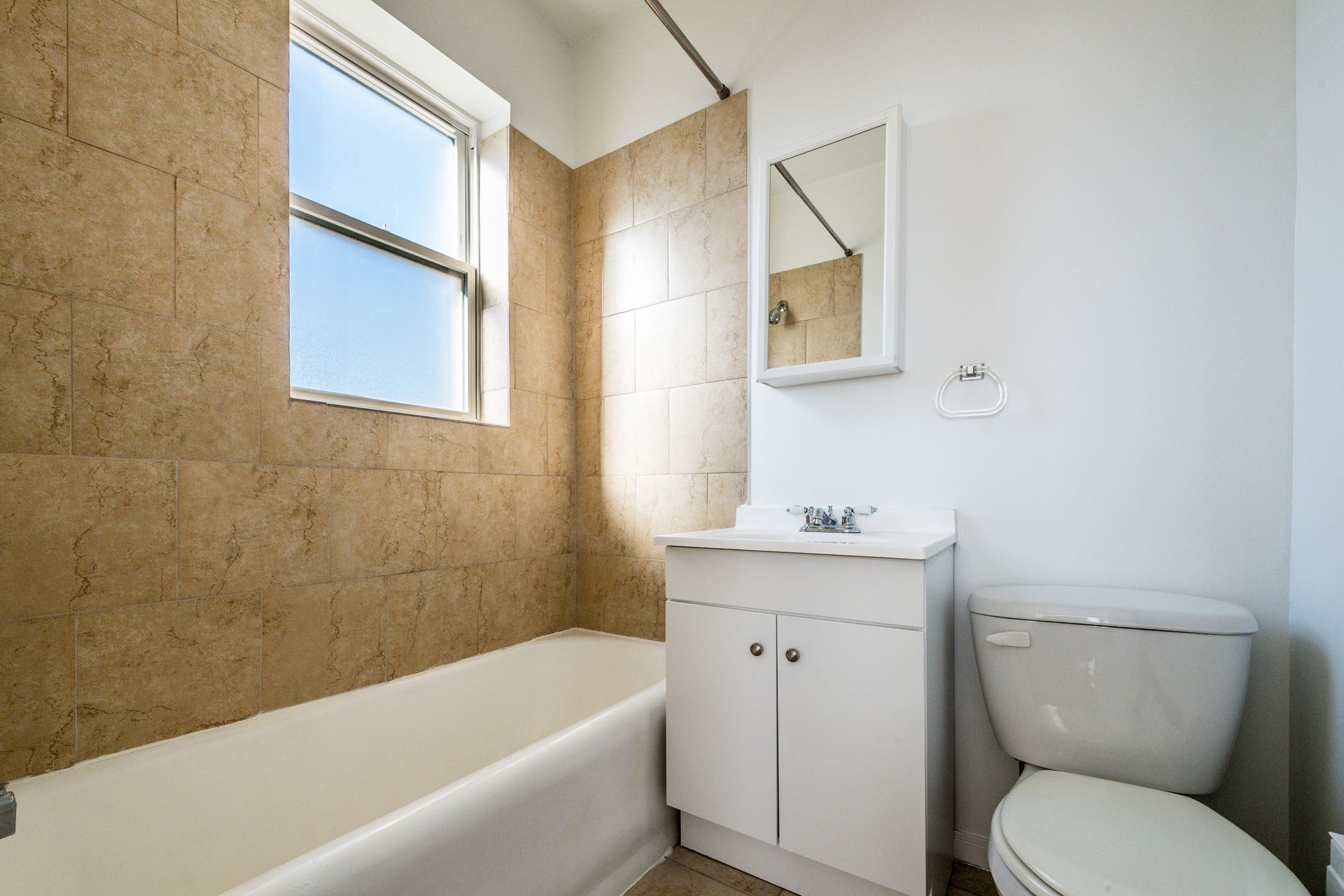 Bathroom with a white bathtub, toilet, and vanity. Beige tiled wall and a window.