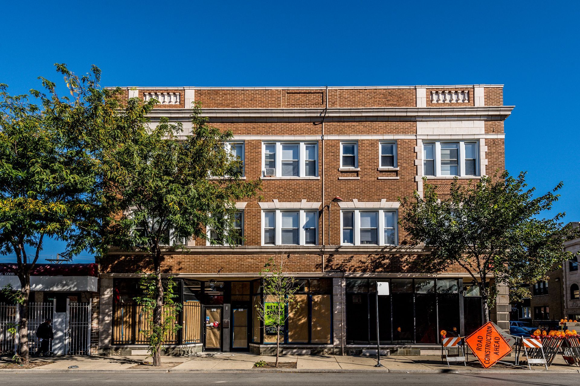 Two-story brick building with boarded up storefronts and windows under a bright blue sky.