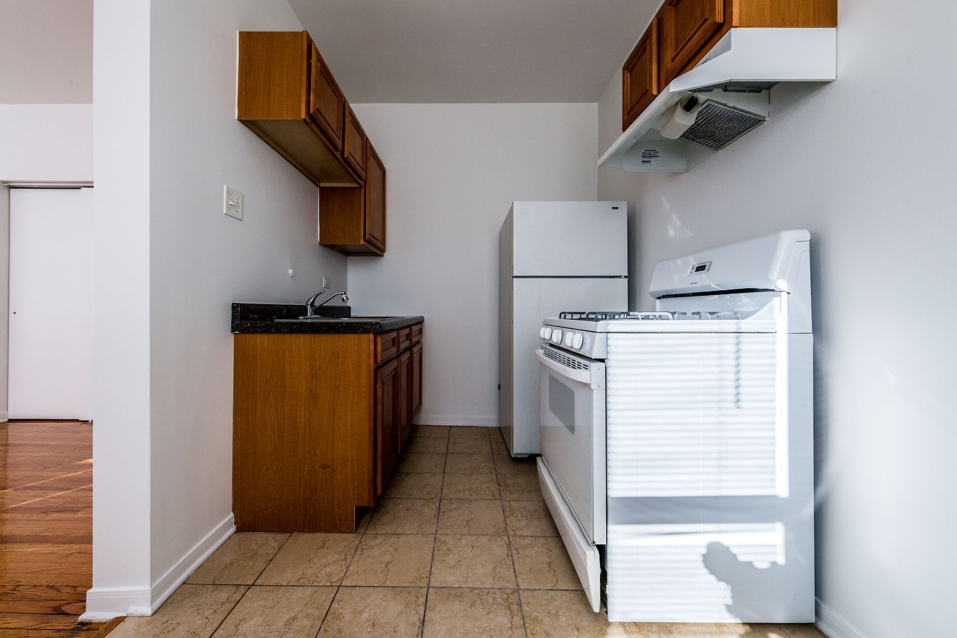 Small kitchen with wooden cabinets, white appliances, and tan tile floor.