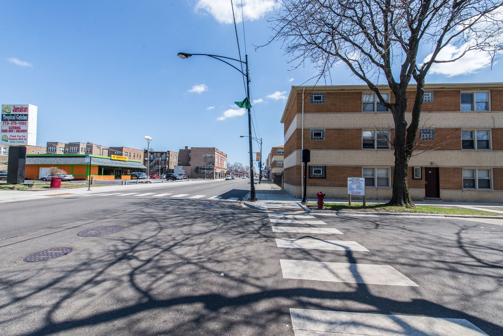 Street view of buildings, blue sky. Shadows of tree across crosswalk.