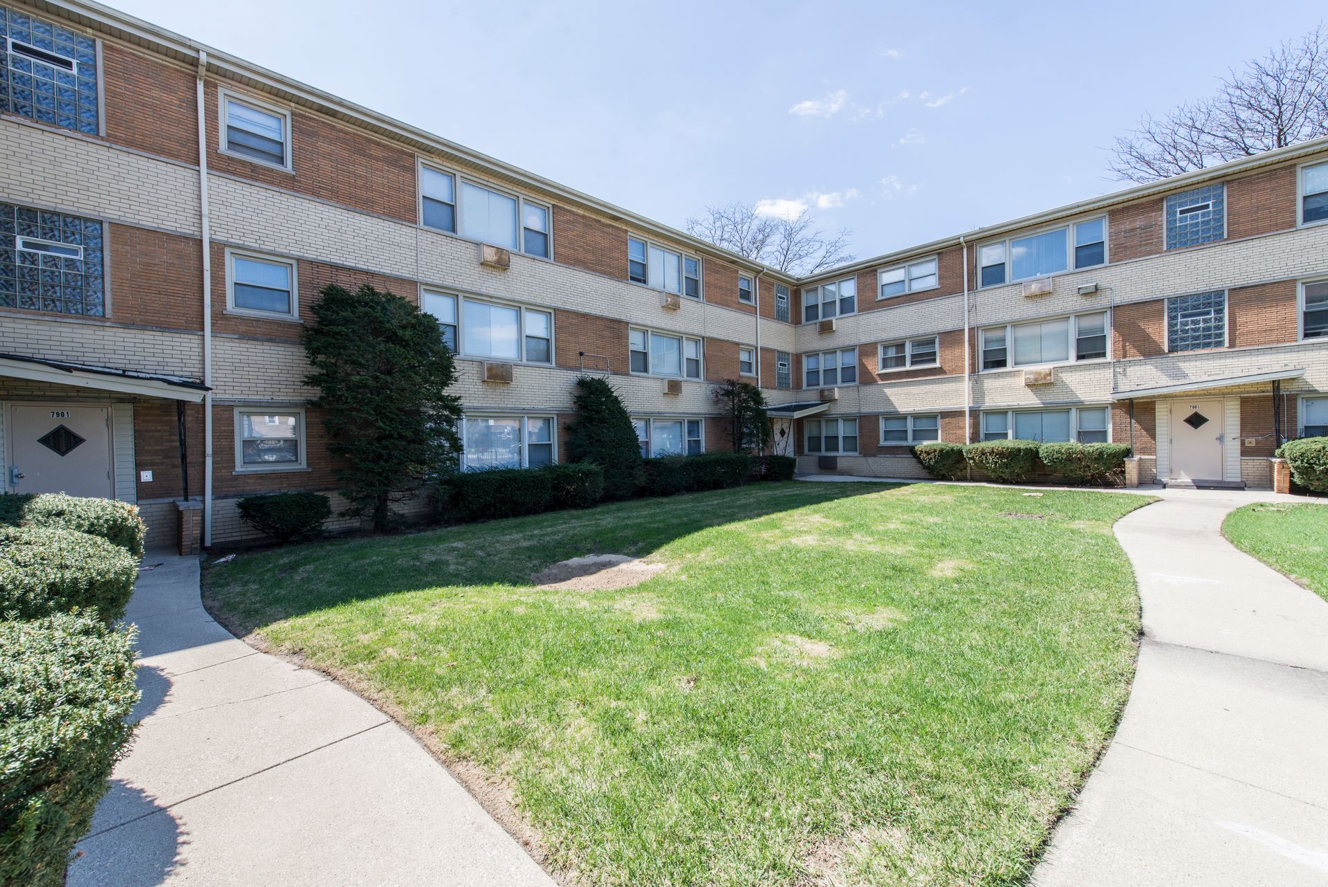 Two-story brick apartment building with a grassy courtyard, walkways, and some trees.
